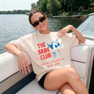 May include: A cream-coloured t-shirt with the text "THE BRIDE CLUB" in red and blue. The shirt features a graphic of a bikini and a cocktail glass. The text "Allison's Bachelorette Miami, FL EST 2020" is also printed on the shirt.