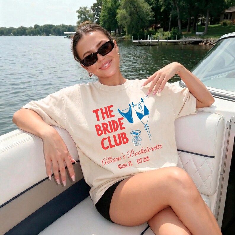 May include: A cream-coloured t-shirt with the text "THE BRIDE CLUB" in red and blue. The shirt features a graphic of a bikini and a cocktail glass. The text "Allison's Bachelorette Miami, FL EST 2020" is also printed on the shirt.