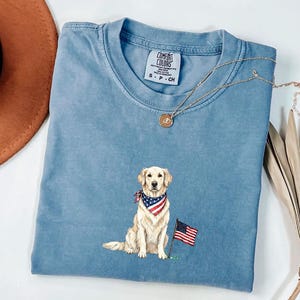 May include: A blue crew neck t-shirt featuring a graphic of a golden retriever dog wearing a red, white, and blue star-spangled bandana. The dog sits next to a small American flag. The shirt has a Comfort Colors brand label at the neckline.