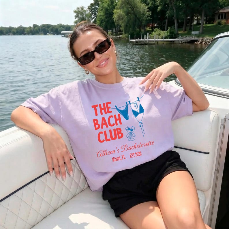 May include: A lilac t-shirt with the text "THE BACH CLUB" in red and blue. The shirt features a bikini graphic and the text "Allison's Bachelorette Miami, FL EST 2020". The person is wearing black shorts and sunglasses.