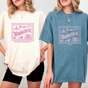 May include: Two short-sleeved t-shirts, one ivory and one blue, each with a pink postage stamp design featuring "Washington D.C." text and landmark illustrations. The ivory shirt has "Postcards from the Bachelorette" text.