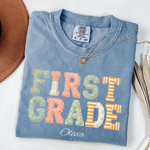 May include: A blue short-sleeved t-shirt featuring the text "FIRST GRADE" in block letters with various floral, polka dot, and striped patterns in shades of peach, green, and white. The name "Olivia" is printed in white script below the text.