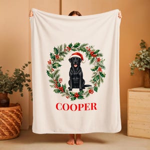May include: A cream-colored fleece blanket featuring a central graphic of a black Labrador dog wearing a red Santa hat, positioned inside a festive wreath of green pine needles and red berries, with the name COOPER printed in red below.