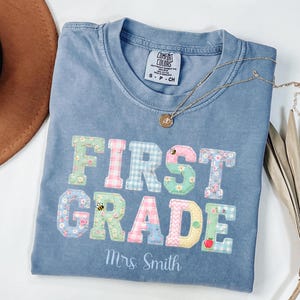 以下が含まれることがあります： パステルカラーの様々な柄が入った大きなブロック体で「FIRST GRADE」と書かれた青い半袖Tシャツ。デザインには小さなハチ、花、リンゴのグラフィックが含まれています。テキストの下には「Mrs. Smith」という名前が白い筆記体でプリントされています。