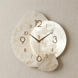 May include: A decorative wall clock with a moon-shaped face and a stone-like frame. The clock features brown hands and numerals, with a glowing moon design in the center. The clock is mounted on a neutral-coloured wall.
