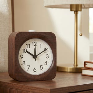 May include: A square, wooden alarm clock with a white face and black hands and numbers. The clock is set to 10:09. The clock sits on a dark brown wooden surface. A gold lamp and a stack of books are in the background.