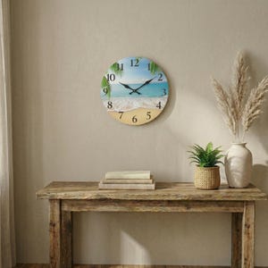 Op de afbeelding: Ronde wandklok met een strandscène. De wijzerplaat toont een blauwe lucht, turquoise water en een zandstrand. Zwarte wijzers geven de tijd aan. De klok is bevestigd aan een beige muur boven een houten tafel.