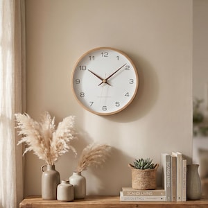 Puede incluir: Un reloj de pared redondo con marco de madera clara y esfera blanca. El reloj tiene números y manecillas negras, con segundero. El reloj está montado en una pared beige. La hora es aproximadamente la 1:10.