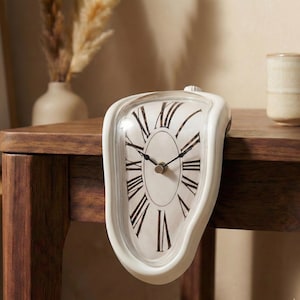 Puede incluir: Un reloj blanco y derretido con números romanos y manecillas negras. El reloj tiene una esfera clara y curvada y un pequeño botón en la parte superior. Descansa sobre una superficie de madera oscura, evocando un tema surrealista.