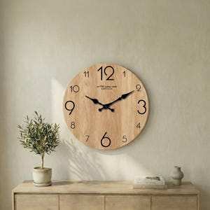 Op de afbeelding: Een ronde houten wandklok met zwarte cijfers en wijzers. De wijzerplaat heeft een licht houtnerfpatroon. De tekst "est 1765 vintage clocks QUARTZ CLOCK" is onder het nummer 12 gedrukt. De klok is aan een beige muur bevestigd.