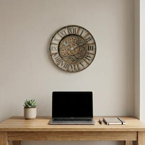 Op de afbeelding: Een ronde wandklok met een metalen frame, Romeinse cijfers en een zichtbaar tandwielmechanisme in het midden. De wijzerplaat heeft een verweerde, neutrale afwerking met donkere wijzers, geplaatst boven een laptop, een kleine vetplant en een notitieboek.