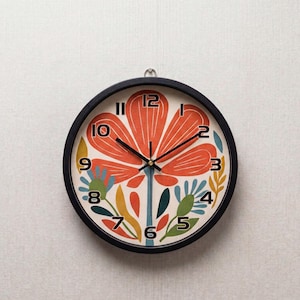 Puede incluir: Un reloj de pared redondo con marco negro. La esfera presenta un gran diseño de flor naranja con números y manecillas negras. Elementos florales adicionales en azul, verde y amarillo rodean la flor, creando una pieza decorativa vibrante.