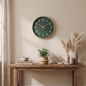 Puede incluir: Un reloj de pared redondo con esfera verde oscuro, números dorados y manecillas doradas. El reloj presenta un marco fino de color dorado y está montado en una pared lisa. El diseño incluye marcadores de hora estándar del uno al doce.