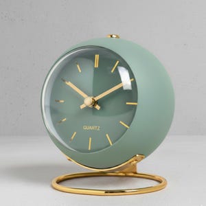 May include: A sage green quartz clock with a spherical design. The clock features gold-colored hands and a gold-colored base. The clock face is visible through a clear, rounded cover. The word "QUARTZ" is printed on the clock face.