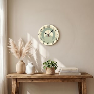 Puede incluir: Un reloj de pared redondo con esfera crema, números negros y manecillas negras. El reloj presenta un diseño verde claro y crema en el centro. El reloj está colgado en una pared beige sobre una mesa de madera. El diámetro del reloj es de unos 25 cm.