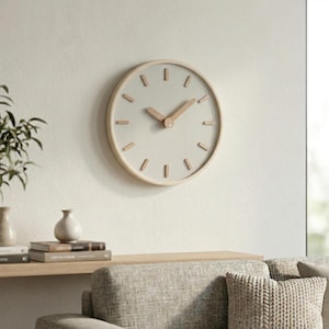 Puede incluir: Un reloj de pared redondo con una esfera gris claro y un marco de madera natural. El reloj tiene marcadores de hora y manecillas de madera. Está montado en una pared blanca, con un entorno interior de tonos neutros en el fondo.