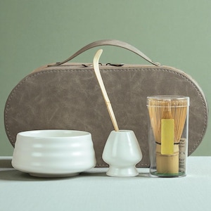 Puede incluir: Un juego de té matcha que incluye un cuenco de cerámica blanco, un batidor de bambú, un soporte para batidor y un estuche de transporte. El batidor está en un recipiente transparente. El juego es para té matcha.