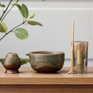 Peut inclure: Un ensemble à thé matcha comprenant un bol en céramique, un fouet et un petit support en céramique. L'ensemble a une palette de couleurs marron et verte et est présenté sur une surface en bois. Un fouet et une cuillère en bambou sont inclus.