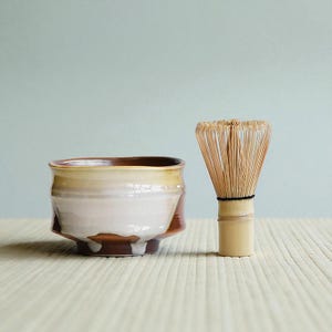 Puede incluir: Un cuenco de matcha de cerámica con borde marrón, esmalte crema y marrón, y un batidor de bambú. El cuenco está sobre una superficie clara con un fondo azul claro. El batidor está hecho de finas púas de bambú.