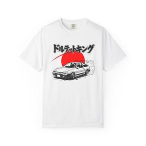 May include: White t-shirt featuring a black and white illustration of a car drifting, with a red sun in the background. Japanese text is above the car. The shirt is a classic crew neck.