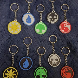 May include: A collection of keychains featuring various symbols in yellow, blue, green, black, and red. Each keychain has a metal ring and chain. The symbols include a sun, water drop, tree, skull, and a flame-like design.