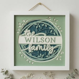 May include: A framed, layered paper art piece with the words "the WILSON family" in white script. The design features a circular arrangement of leaves in shades of green and blue, set against a light green background. The frame is white, and a rope is used for hanging.