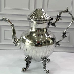 May include: A polished silver teapot featuring a rounded body with vertical fluted detailing at the base, a curved spout, an ornate handle, and a matching lid. The teapot stands on three decorative feet and has a highly reflective metallic surface.