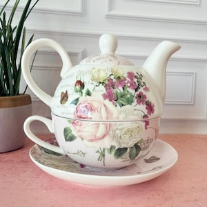 May include: A white ceramic tea set for one, featuring a teapot that stacks onto a teacup, resting on a matching saucer. The set is decorated with a vintage-style floral print of pink and white roses, small purple flowers, and a butterfly.