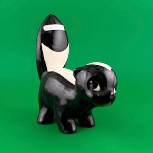 May include: A glossy ceramic skunk figurine featuring a black body with a white stripe running from the top of its head down its back and onto its upright tail. The skunk has a small white sticker on its tail with the text PREM printed in black.