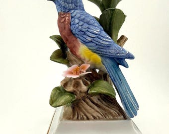 May include: A porcelain figurine of a bluebird with a reddish-brown chest and yellow belly, perched on a brown tree stump with green leaves and a small pink flower. The bird is mounted on a white rectangular base with gold trim and scrollwork.