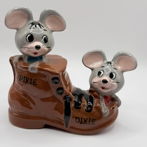 May include: A ceramic figurine featuring two cartoon mice in a brown boot. One mouse wears a blue bow tie, the other a red bow tie. The boot is inscribed with the words "PIXIE" and "DIXIE".