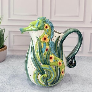 May include: A ceramic pitcher shaped like a seahorse with a white base, featuring hand-painted green and blue seaweed patterns, yellow flowers with red centers, and a curved handle. The surface is decorated with small brown dots and a signature.