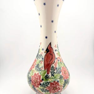 May include: A ceramic vase with a cream-colored base featuring a hand-painted red cardinal bird, green holly leaves, and pink poinsettia flowers. The neck is decorated with small blue star-like patterns and a dark blue rim at the top and base.