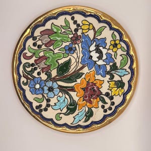 May include: A round, decorative plate with a cream-colored background and a floral design. The flowers are in shades of blue, orange, and red, with green leaves. The plate has a gold rim and a blue scalloped border. The plate is approximately 8 inches in diameter.