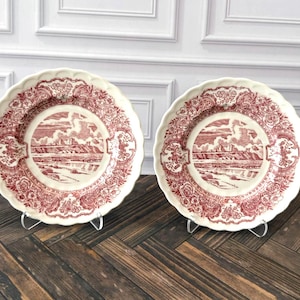 May include: Two round ceramic plates with scalloped edges, featuring a deep red transferware pattern on a cream background. The central design depicts a landscape with mountains and clouds, surrounded by a border of intricate floral motifs.