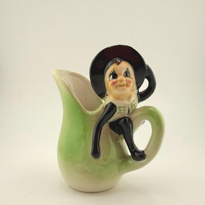 May include: A vintage ceramic creamer pitcher with a whimsical design. The pitcher is light green with a cream-colored interior. A cartoon figure with a black hat and legs is perched on the pitcher's handle. The figure has a smiling face and is wearing a plaid top.