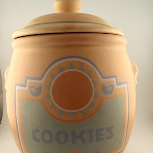 May include: A tan ceramic cookie jar with a lid and handles. The jar features a decorative label with the word "COOKIES" in blue letters. The label has a sun design and is outlined in blue and white.