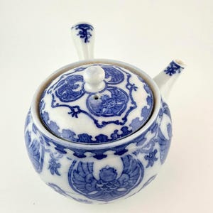 May include: A white ceramic teapot with a side handle and a matching lid, decorated with intricate blue patterns featuring stylized bird motifs and floral elements. The teapot has a rounded body and a small spout, showcasing a classic blue and white design.