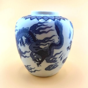 May include: A white ceramic jar featuring a blue dragon motif with intricate line work. The vessel has a rounded body, a short neck, and a decorative geometric pattern encircling the rim. The dragon design is depicted in a dark blue pigment.