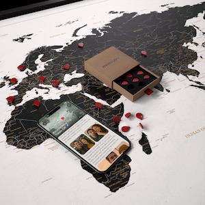 Può includere: Una mappa del mondo in bianco e nero con dettagli dorati, cosparsa di spilli rossi a forma di cuore. Uno smartphone mostra foto di viaggio e testo. Una scatola con la scritta "memories" contiene altri spilli a forma di cuore.