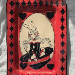 Puede incluir: Una bandeja rectangular de cerámica con un borde rojo brillante y un patrón de diamantes negros. El centro muestra la ilustración de un personaje con cabello largo blanco, orejas de conejo y ojos rojos, vistiendo un atuendo negro.