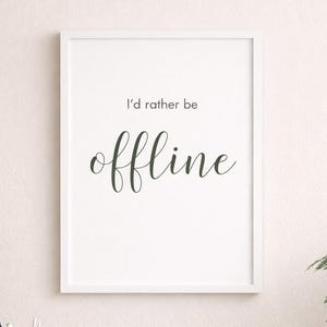 May include: A framed print with the text "I'd rather be offline" in a white frame. The print is on a desk with a white mug, a notebook, a phone, and a pencil holder. A white chair is in front of the desk.
