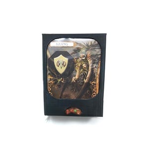 May include: Black box with a fantasy art print featuring a monster and a shield with a gold emblem. The word "KRANG" is printed at the top. The box has a small, decorative, brown and green button.