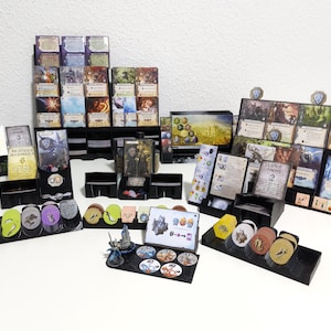 Könnte beinhalten: Eine Sammlung von Brettspielkomponenten, darunter Karten, Spielsteine und Miniaturfiguren. Die Karten sind in schwarzen Haltern gestapelt, und die Spielsteine sind in verschiedenen Farben. Die Spielteile sind auf einer weißen Oberfläche angeordnet.