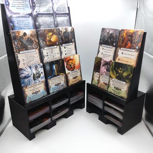 Könnte beinhalten: Zwei schwarze Kartenhalter aus Holz, jeweils mit mehreren Fächern zur Organisation von Sammelkarten. Die Halter zeigen verschiedene Karten mit Fantasy-Kunst und deutschem Text. Die Halter sind für eine einfache Anzeige und den Zugriff abgewinkelt.