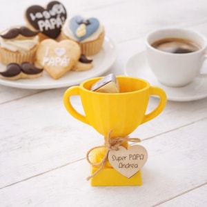 Può includere: Un trofeo giallo con un'etichetta a forma di cuore con la scritta "Super PAPA Andrea". La coppa è legata con spago e contiene piccoli oggetti. Cupcake, biscotti e una tazza di caffè sono sullo sfondo.