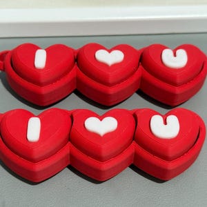 Può includere: Oggetti rossi a forma di cuore con lettere bianche che compongono "I U". Ogni cuore ha una piccola maniglia laterale. Le lettere sono bianche e i cuori sono rossi.
