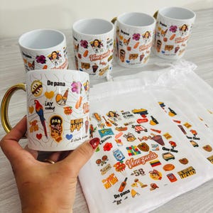 May include: White ceramic mugs with gold handles, decorated with colorful Venezuelan-themed stickers and illustrations. The mugs feature images of a woman, food, and phrases like "De pana" and "Venezuela". A matching white cloth with similar designs is also present.