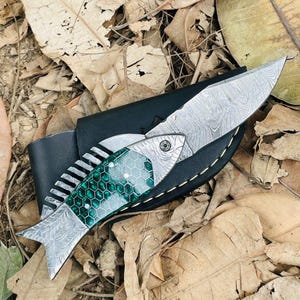 May include: A decorative knife with a fish-shaped handle and a patterned blade. The handle features a green and silver design, and the blade has a textured pattern. The knife is resting in a black leather sheath.