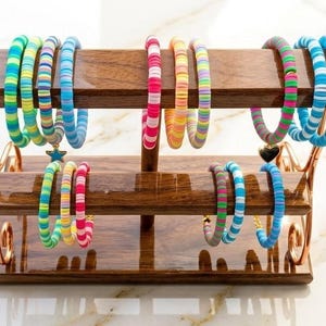 May include: A two-tiered wooden jewelry display stand with a polished finish and copper-toned metal supports, holding multiple colorful clay bead bracelets in various patterns, including blue, green, pink, yellow, and purple hues.